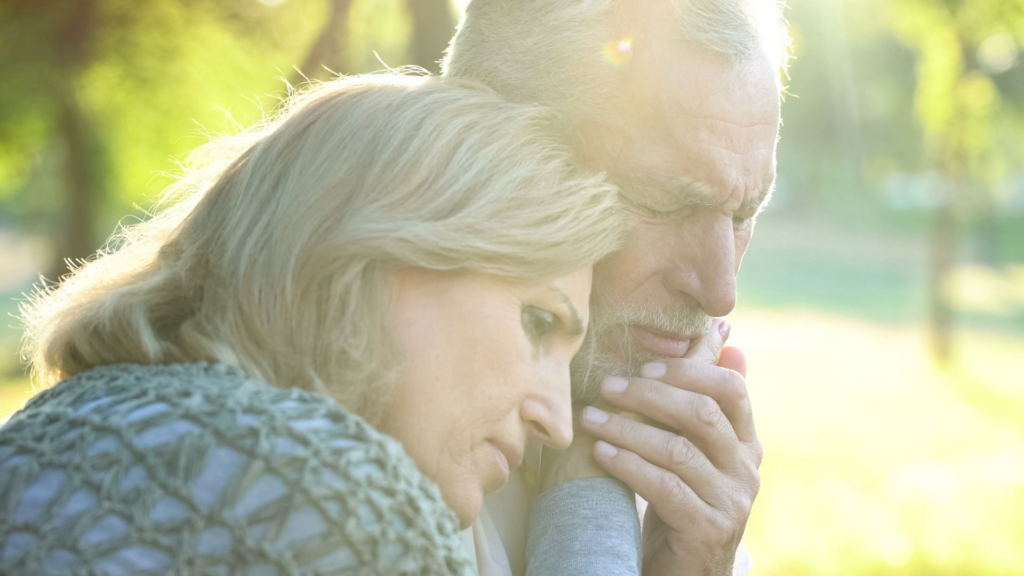 An older couple, close together in what looks like a park. The woman has her head on the man's shoulder. They both look sad.