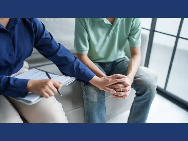 Two people sit in a clinical setting. One holds a clipboard and reaches out to hold the other person's hands, showing support and care.